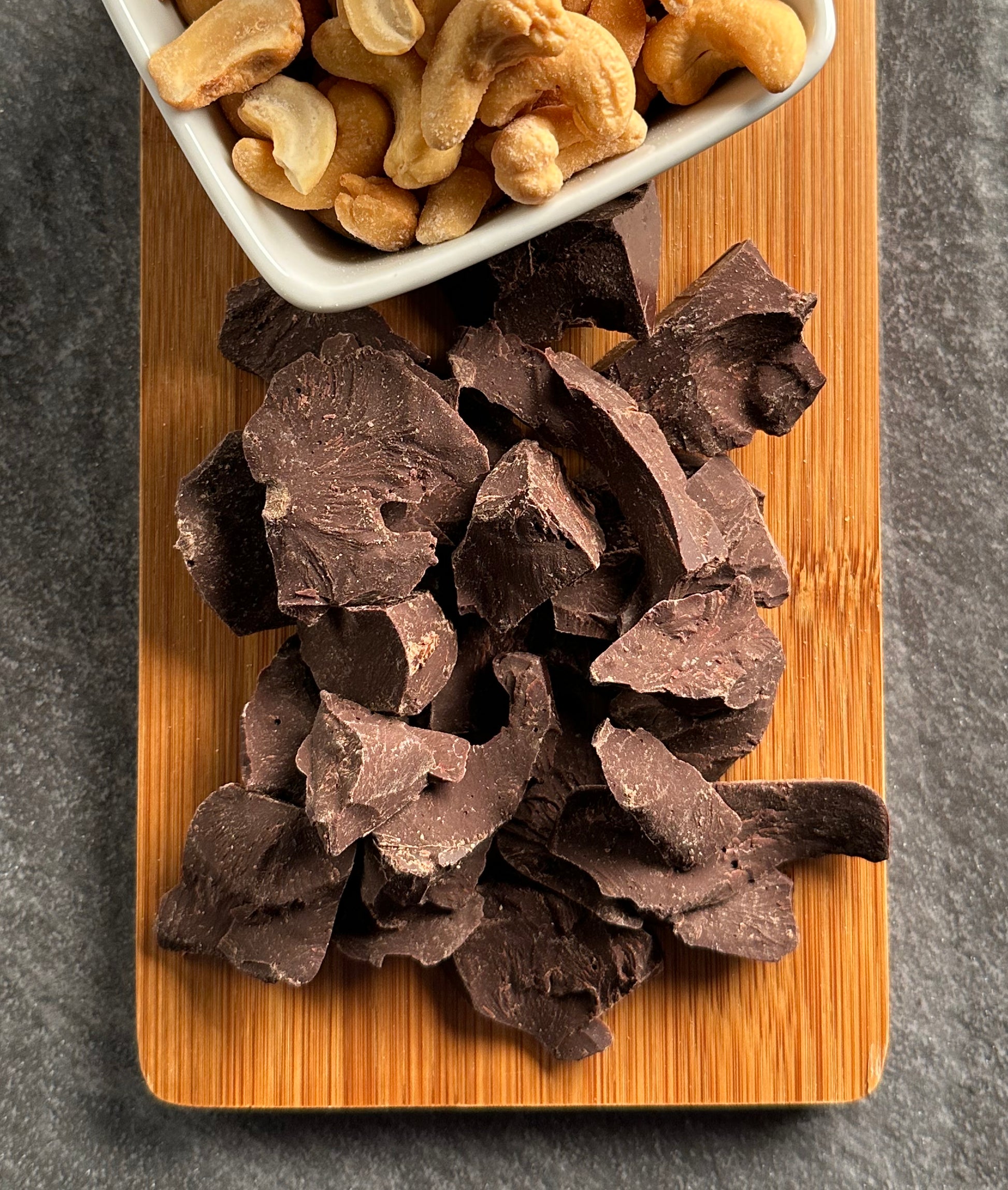 A dessert board displays The Chocolate Butcher’s SemiSweet Dark Chocolate — Roughly Chopped at the center and a small white dish of cashew nuts above, set on a wooden board against a dark grey surface for an indulgent treat.