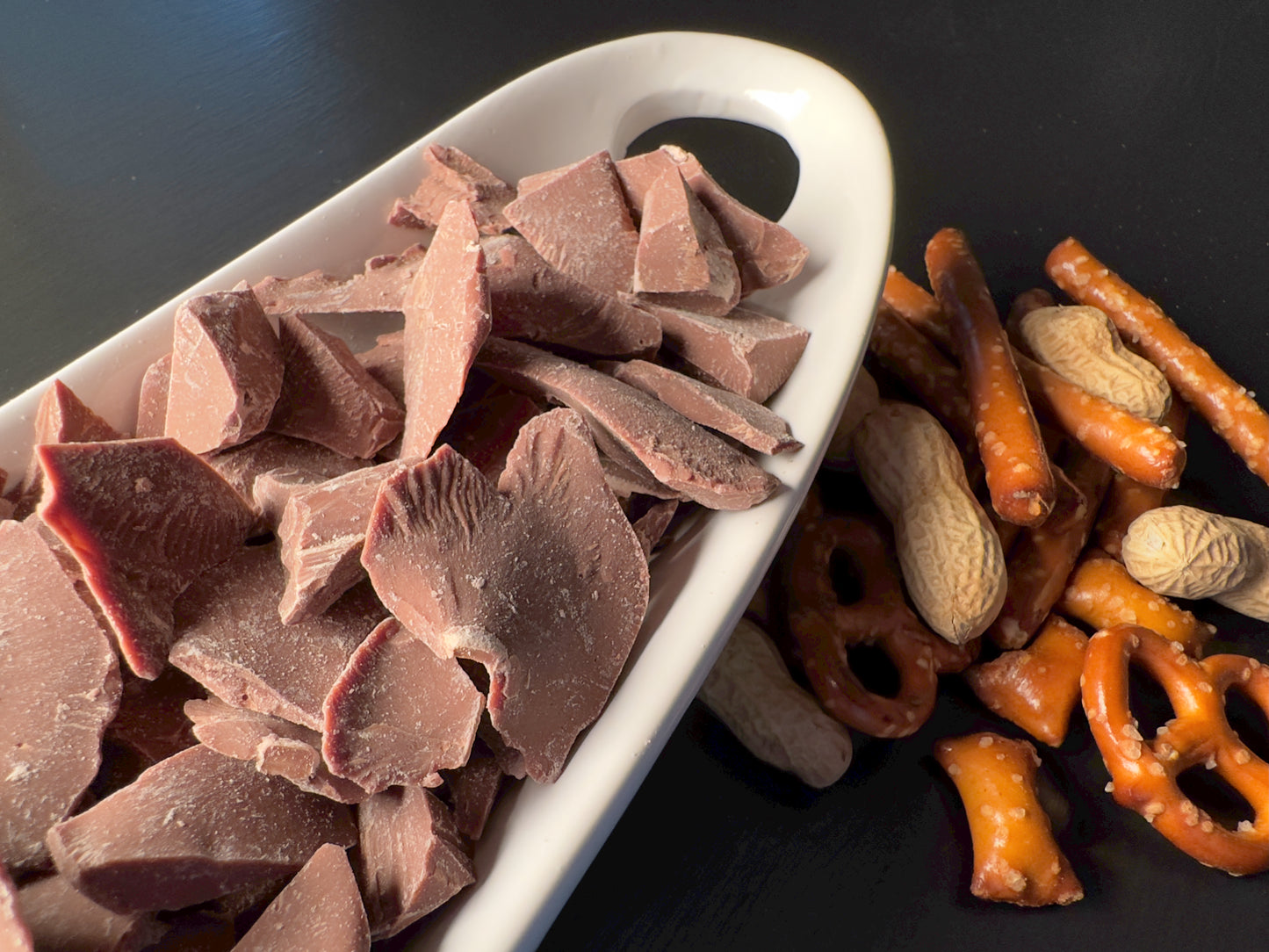 A white dish holds The Chocolate Butcher’s Milk Chocolate — Roughly Chopped, set beside salted pretzel twists, pretzel sticks, and whole peanuts—an ideal addition to any dessert board.